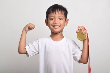 Asyalı çocuk altın madalyasını tutuyor ve beyaz arka planda izole edilmiş kol kaslarını gösteriyor. Çocuk zaferini kutluyor. İlk olarak spor kupası.