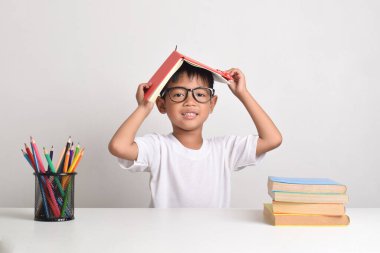 Kafasında bir kitapla beyaz bir arka planda okuyan Asyalı bir çocuğun portresi. Çocuk ev ödevini bitirdikten sonra mutlu bir şekilde gülümser.