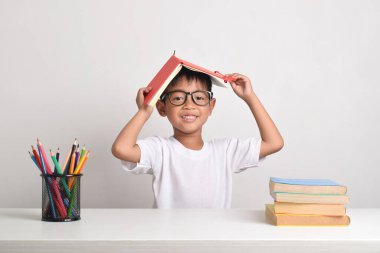 Kafasında bir kitapla beyaz bir arka planda okuyan Asyalı bir çocuğun portresi. Çocuk ev ödevini bitirdikten sonra mutlu bir şekilde gülümser.