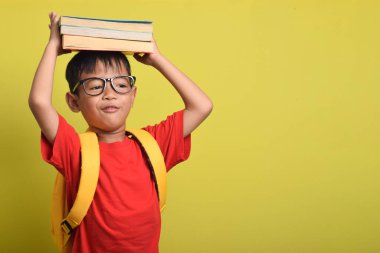 Kafasında bir kitap ve sarı arka planda izole edilmiş bir çanta olan sevimli ve neşeli Asyalı çocuk, uzayı kopyala. Mutlu çocuk. Okula dön..