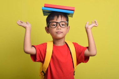 Okula dönelim. Sırt çantalı ve kafasında sarı arka planda izole edilmiş kitaplar taşıyan neşeli Asyalı çocuk. Çocuk çalışmaya hazır.