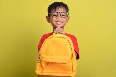 Sarı sırt çantalı, gülümseyen ve sarı arka planda izole edilmiş kameraya bakan iyimser Asyalı çocuk. Mutlu okul çocuğu. Okula geri dön.