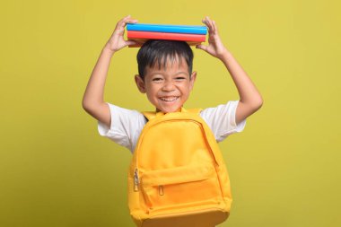 Okula dönelim. Sırt çantalı ve kafasında sarı arka planda izole edilmiş kitaplar taşıyan neşeli Asyalı çocuk. Çocuk çalışmaya hazır.