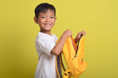 Sarı arka planda, sırt çantasının açık duran neşeli Asyalı çocuğun portresi. Okul için hazırlık. Kameraya bakıyorum. Okul konseptine dönüş