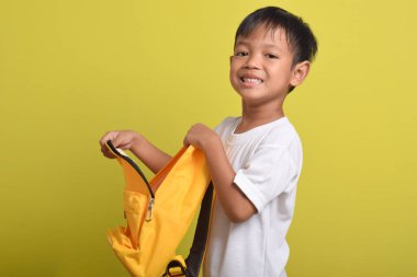 Sarı arka planda, sırt çantasının açık duran neşeli Asyalı çocuğun portresi. Okul için hazırlık. Kameraya bakıyorum. Okul konseptine dönüş