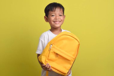 Sarı arka planda izole edilmiş okul çantası tutan mutlu bir Asyalı çocuk. Çalışmaya hazırlanan sevimli bir çocuk. Okul konseptine dönüş