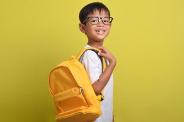 Sırt çantalı Asyalı çocuk sarı arka planda izole edilmiş kameraya gülümsüyor. Şirin bir çocuk ders çalışmaya hazırlanıyor. Okula Dönüş