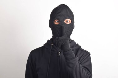 Siyah ceketli ve maskeli bir hırsız parmaklarını dudaklarına dayayıp, beyaz arka planda izole edilmiş sessiz bir işaret yapıyor. Hırsızlık ve suç kavramı