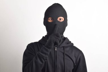 Siyah ceketli ve maskeli bir hırsız parmaklarını dudaklarına dayayıp, beyaz arka planda izole edilmiş sessiz bir işaret yapıyor. Hırsızlık ve suç kavramı