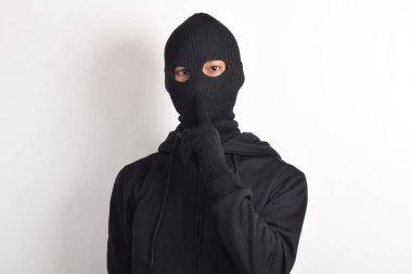 Siyah ceketli ve maskeli bir hırsız parmaklarını dudaklarına dayayıp, beyaz arka planda izole edilmiş sessiz bir işaret yapıyor. Hırsızlık ve suç kavramı