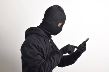 Siyah kapüşonlu ve maskeli gizemli bir adam cep telefonunda hackerlık yapıyor, akıllı telefon tutuyor, beyaz arka planda izole edilmiş..
