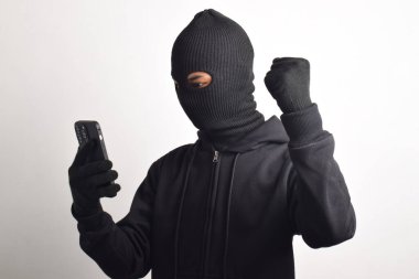 Siyah kapüşonlu ve maskeli gizemli adam akıllı telefondan hackleme faaliyetinde başarılı ve mutlu bir ifadeyle beyaz arka planda izole edilmiş..