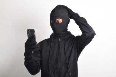 Siyah kapüşonlu ve maskeli gizemli bir adam akıllı telefondan bilgisayar korsanlığı yapıyor, kafası karışmış, beyaz arka planda izole edilmiş..