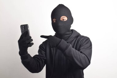 Siyah kapüşonlu ve maskeli gizemli adam akıllı telefondan hackleme aktivitesi yapıyor, akıllı telefonu işaret ediyor, beyaz arka planda izole edilmiş..