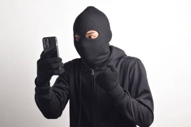 Siyah kapüşonlu ve maskeli gizemli adam akıllı telefondan hackleme faaliyetinde başarılı ve mutlu bir ifadeyle beyaz arka planda izole edilmiş..