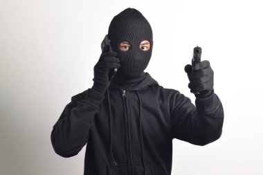 Siyah kapüşonlu ve maskeli gizemli adam telefon görüşmesinde sahtekarlık yapıyor, kameraya silah doğrultuyor, beyaz arka planda izole edilmiş..