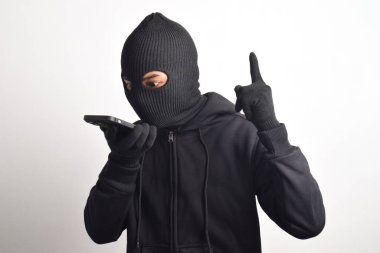 Siyah kapüşonlu ve maskeli gizemli bir adam. Akıllı telefondan para sızdırıyor, beyaz arka planda izole edilmiş..