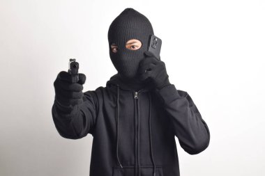 Siyah kapüşonlu ve maskeli gizemli adam telefon görüşmesinde sahtekarlık yapıyor, kameraya silah doğrultuyor, beyaz arka planda izole edilmiş..