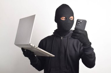 Siyah kapüşonlu ve maskeli gizemli adam bilgisayar ve akıllı telefon hackleme faaliyetleri yapıyor, kişisel bilgileri çalıyor, beyaz arka planda izole edilmiş..