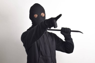 Siyah ceketli ve maskeli bir hırsız levye tutuyor, kameraya bakıyor, beyaz arka planda izole edilmiş..