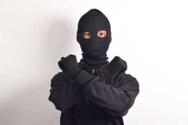 Siyah kapüşonlu ve maskeli gizemli bir adam haç işareti yapıyor ve beyaz arka planda dışlanmış bir yüz ifadesi sergiliyor..