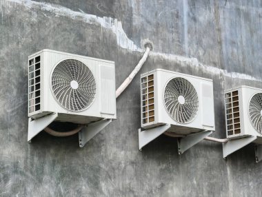 Many white outdoor air conditioning units mounted on a grey, textured wall, with visible pipes.