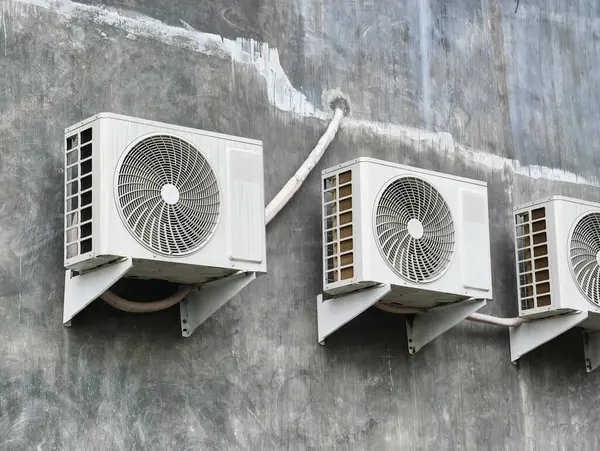 Many white outdoor air conditioning units mounted on a grey, textured wall, with visible pipes.