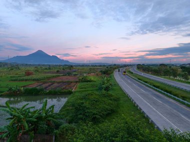 Gün batımında yemyeşil tarlaları ve küçük bir su kütlesi olan uzak bir dağa doğru uzanan güzel bir otoyol..