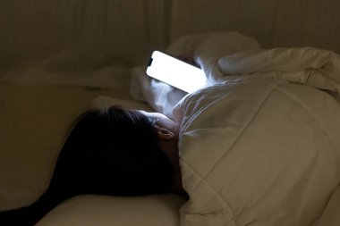 Woman looking at her cellphone under the white blanket, the girl lying and rolling in the bed at night before sleep