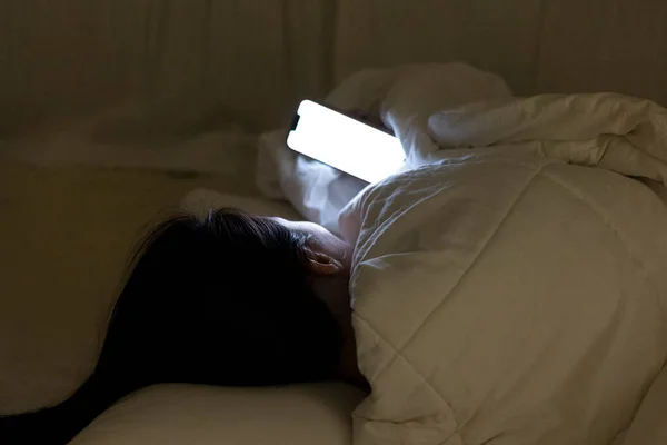 Woman looking at her cellphone under the white blanket, the girl lying and rolling in the bed at night before sleep