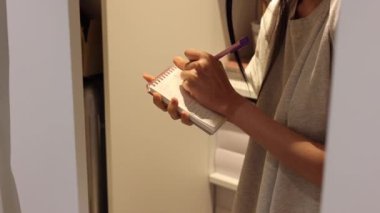 Woman is writing down something on a notebook, worker is taking inventory in a warehouse