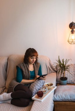 Dikey, kadın, orta yaşlı, beyaz, kısa saçlı, çekici, günlük kıyafetler giyen, kitap okuyan, aşılanmış ve fındıklı. Pencerenin yanındaki kanepede oturuyor..