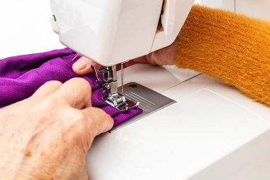 Son derece yatay yakın çekim fotoğrafı, yetişkin beyaz kadın terzinin elleri elle terzilik işi yapıyor. Mor bir kumaş dikiyorum, beyaz bir masada dikiş makinesine..