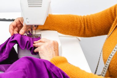 Son derece yatay yakın çekim fotoğrafı, yetişkin beyaz kadın terzinin elleri elle terzilik işi yapıyor. Mor bir kumaş dikiyorum, beyaz bir masada dikiş makinesine..