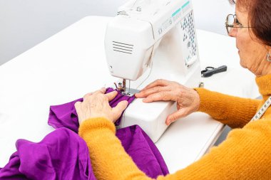 Orta boy yatay fotoğraf, yetişkin beyaz kadın terzinin elleri elle terzilik yapıyor. Mor bir kumaş dikiyorum, beyaz bir masada dikiş makinesine..