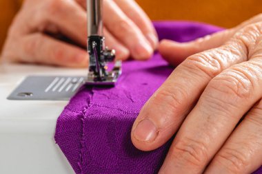 Son derece yakın fotoğraf, yatay eller olgun yetişkin kadın terzi, beyaz. Turuncu kazak, dikiş makinesiyle dikiş mor bir kumaş..