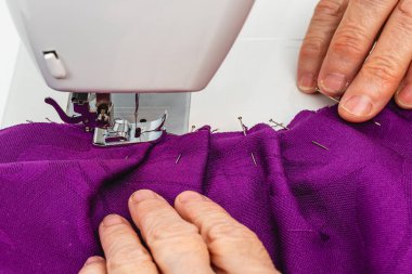 Son derece yatay yakın çekim fotoğrafı, yetişkin beyaz kadın terzinin elleri elle terzilik işi yapıyor. Mor bir kumaş dikiyorum, beyaz bir masada dikiş makinesine..