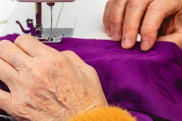 Extreme close up photo, horizontal hands mature adult woman seamstress, Caucasian. with orange sweater, sewing with the sewing machine a purple fabric.