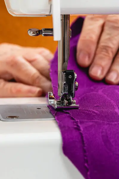 Extreme close up photo, vertical hands mature adult female seamstress, Caucasian. with orange sweater, sewing with the sewing machine a purple fabric.