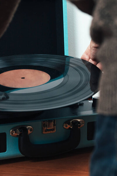 Vertical close up photo, senior adult male, Caucasian, back turned, dressed in brown wool sweater. Placing a music vinyl on a record player in the shape of a blue suitcase. Concept hobbies, art.