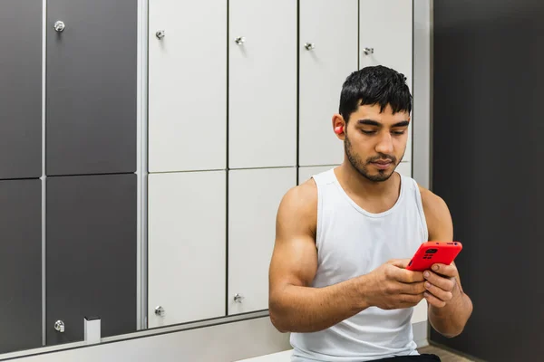 Horizontal photo man young adult mixed race, in the gym locker room, sitting with the cell phone in his hands and a red earphone in his ear. Copy space. Sports, technology concept.