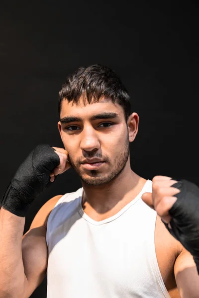Vertical photo portrait of man young adult mixed race boxer looking at camera with fists with black boxing bandages raised, on black background. Sports, recreation concept.