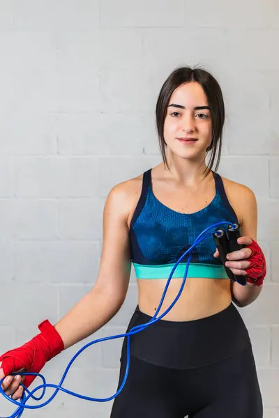 Vertical photo pretty Caucasian teen girl, dressed in sports clothes, with red bandages on her hands and a blue rope. In a boxing gym on white brick copy space background. Concept sports, recreation.