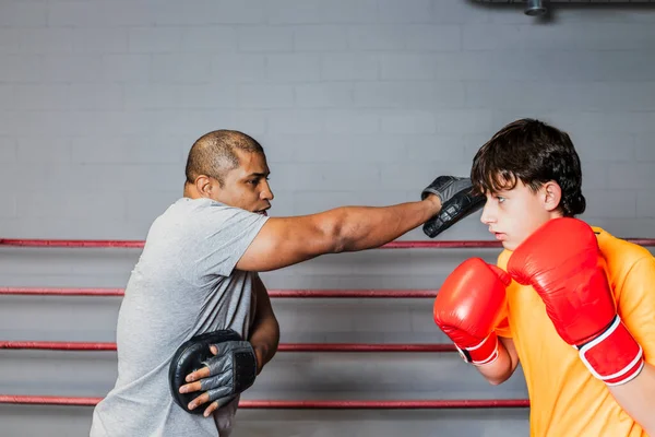 Horizontal photo caucasian teen boy with orange t-shirt and red boxing gloves, in the ring, training with his mid adult black boxing teacher. Concept sports, recreation.