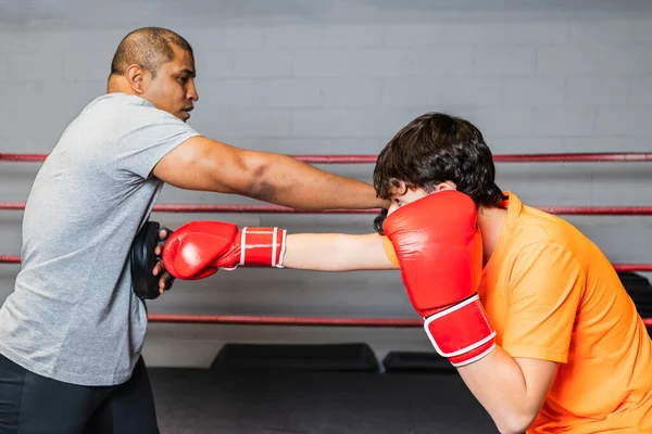 Horizontal photo caucasian teen boy with red boxing gloves and orange t-shirt, training in the ring of a boxing school, with his mid adult black teacher. Concept sports, recreation.