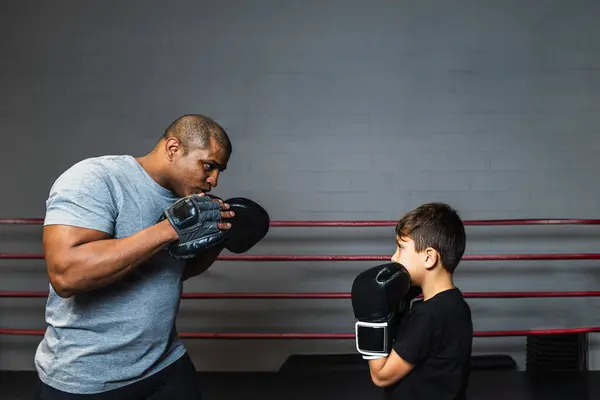 Horizontal photo boy elementary, dressed in t-shirt and black boxing gloves in the ring with his black mid adult boxing trainer, concept sport, recreation.