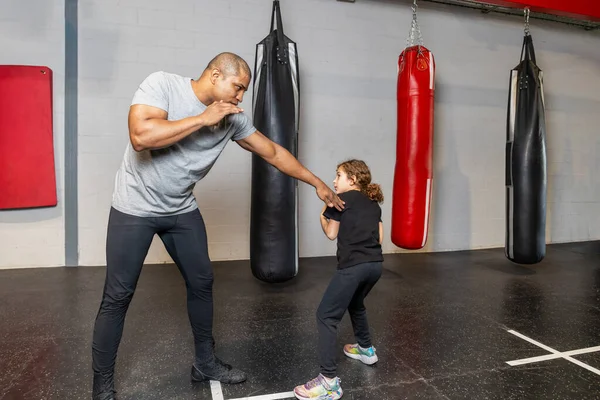 Horizontal photo elementary girl, dressed in tights and black t-shirt, warming up with her black mid adult teacher at a boxing school. Sport, recreation concept.
