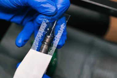 Yatay fotoğraf. Mavi eldivenli bir dövme sanatçısının ellerini yakından görmek. Kullanmak için yeni bir iğneyle dövme makinesini dikkatlice hazırlamak. Uzayı kopyala İş dünyası, sanat.