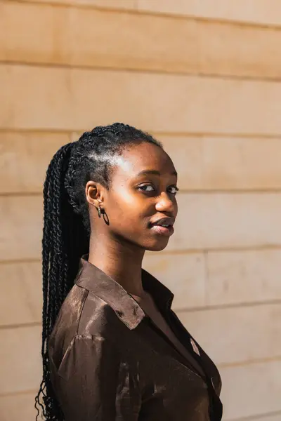 Vertical photo close-up of a confident woman african with long black braids and a brown satin shirt, standing against a beige wall. People concept.