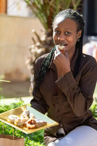 Vertical photo a woman african with braided hair delights in tasting a homemade sweet treat, sitting in a sunlit garden with a plate of arabic pastries. Food and culture concept.